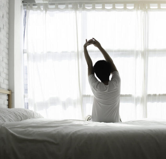 Person dehnt sich morgens im Bett vor hellem Fenster