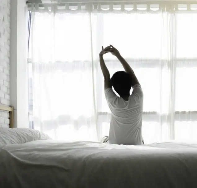 Person dehnt sich morgens im Bett vor hellem Fenster