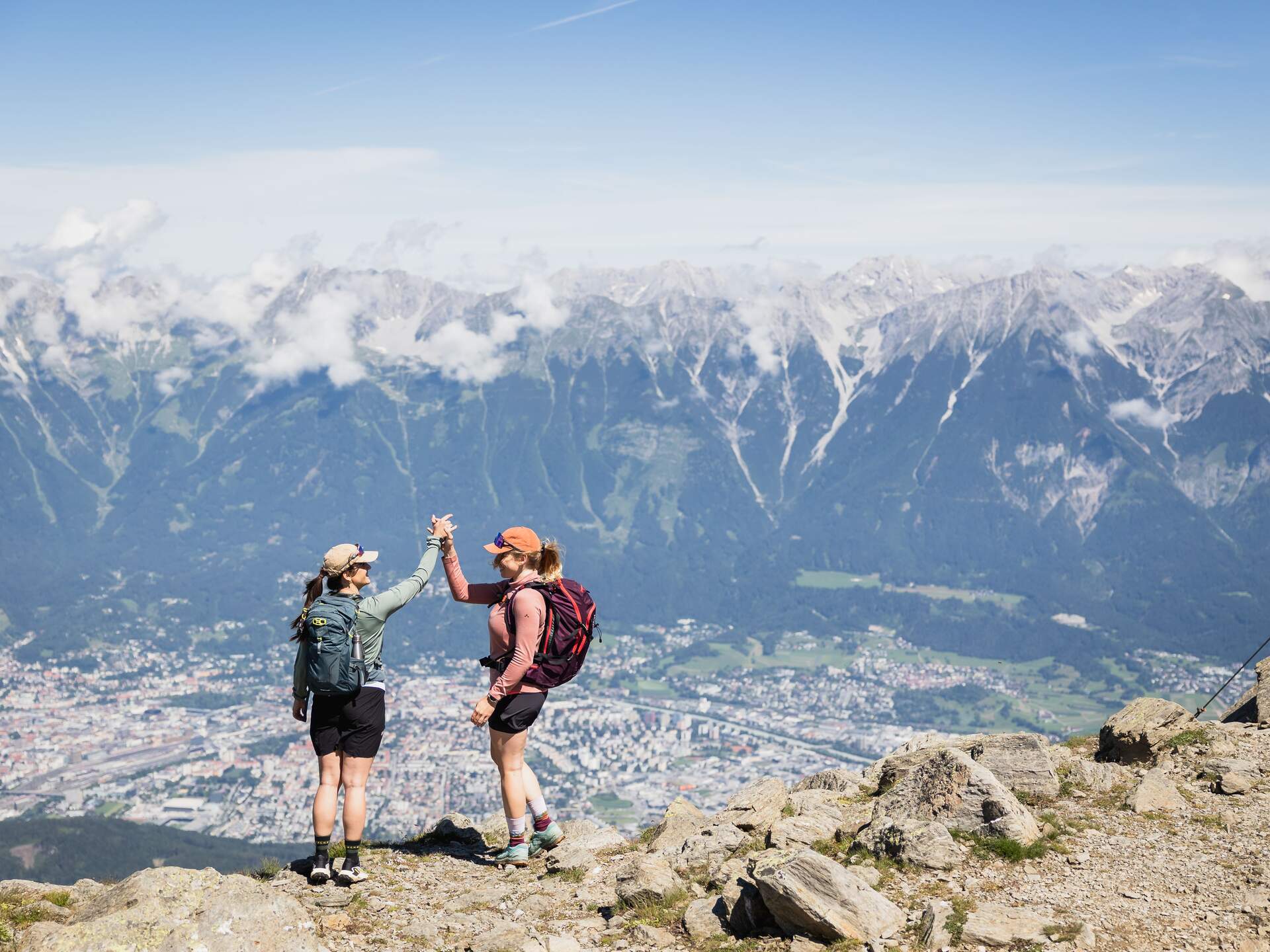 Two hikers high-five each other on a mountain peak overlooking a city and snow-capped mountains | © Jonas Schwarzwälder Photography