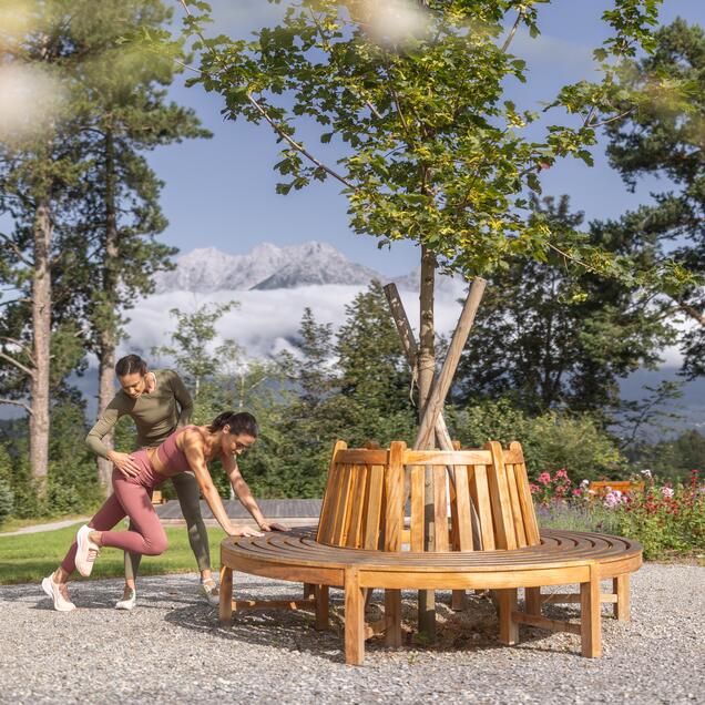 Young woman trains with personal trainer on round wooden bench in park against mountain backdrop