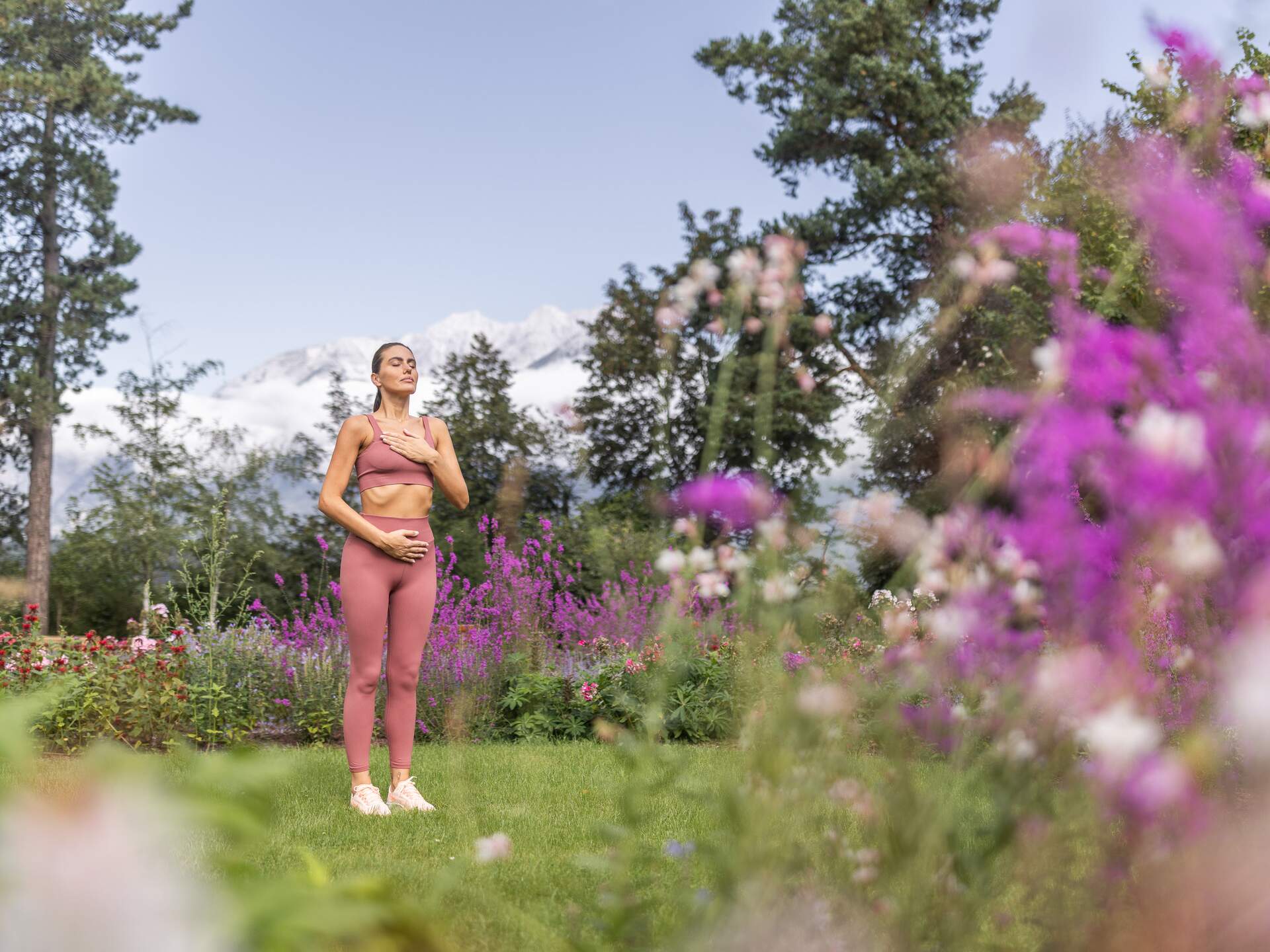 Frau in Sportkleidung meditiert entspannt im Garten mit Blumen und Bäumen vor Bergkulisse