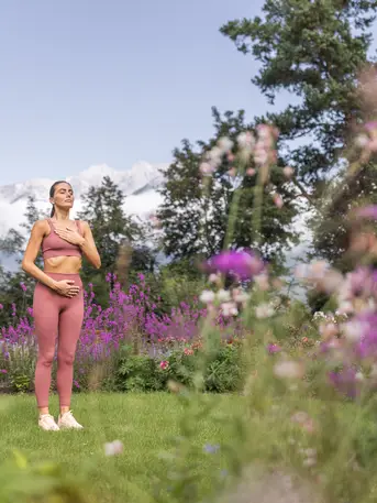 Woman in sportswear meditates relaxed in the garden with flowers and trees in front of mountain backdrop