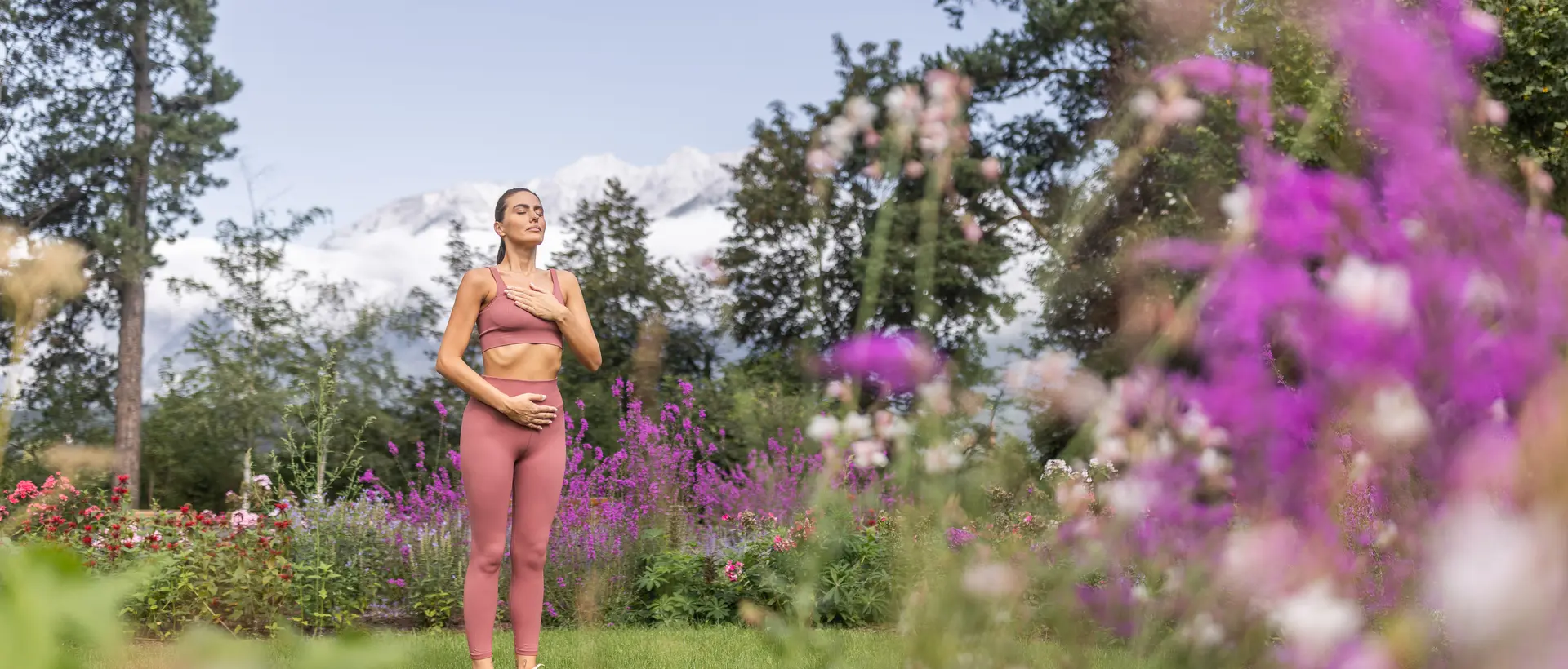 Frau in Sportkleidung meditiert entspannt im Garten mit Blumen und Bäumen vor Bergkulisse